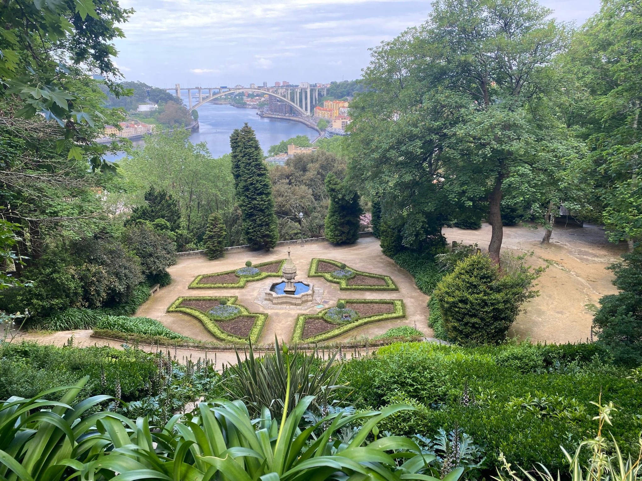 Ornate garden with fountain, surrounded by trees, overlooking a river and arched bridge in the background.