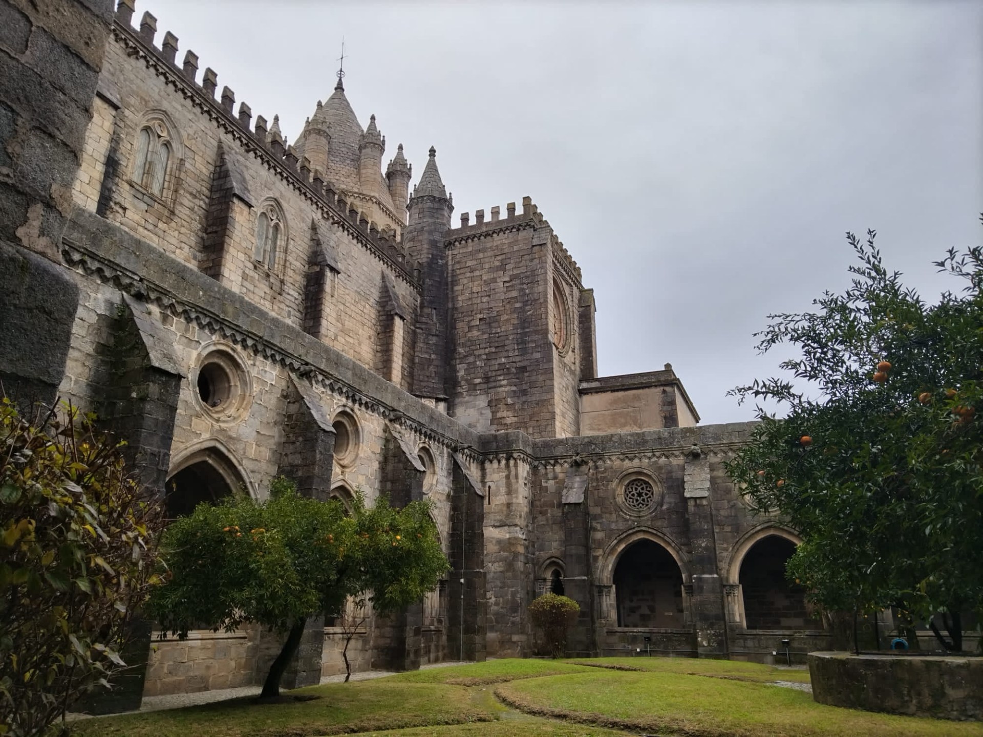 Gothic cathedral courtyard with arched windows, stone walls, and orange trees on a cloudy day.