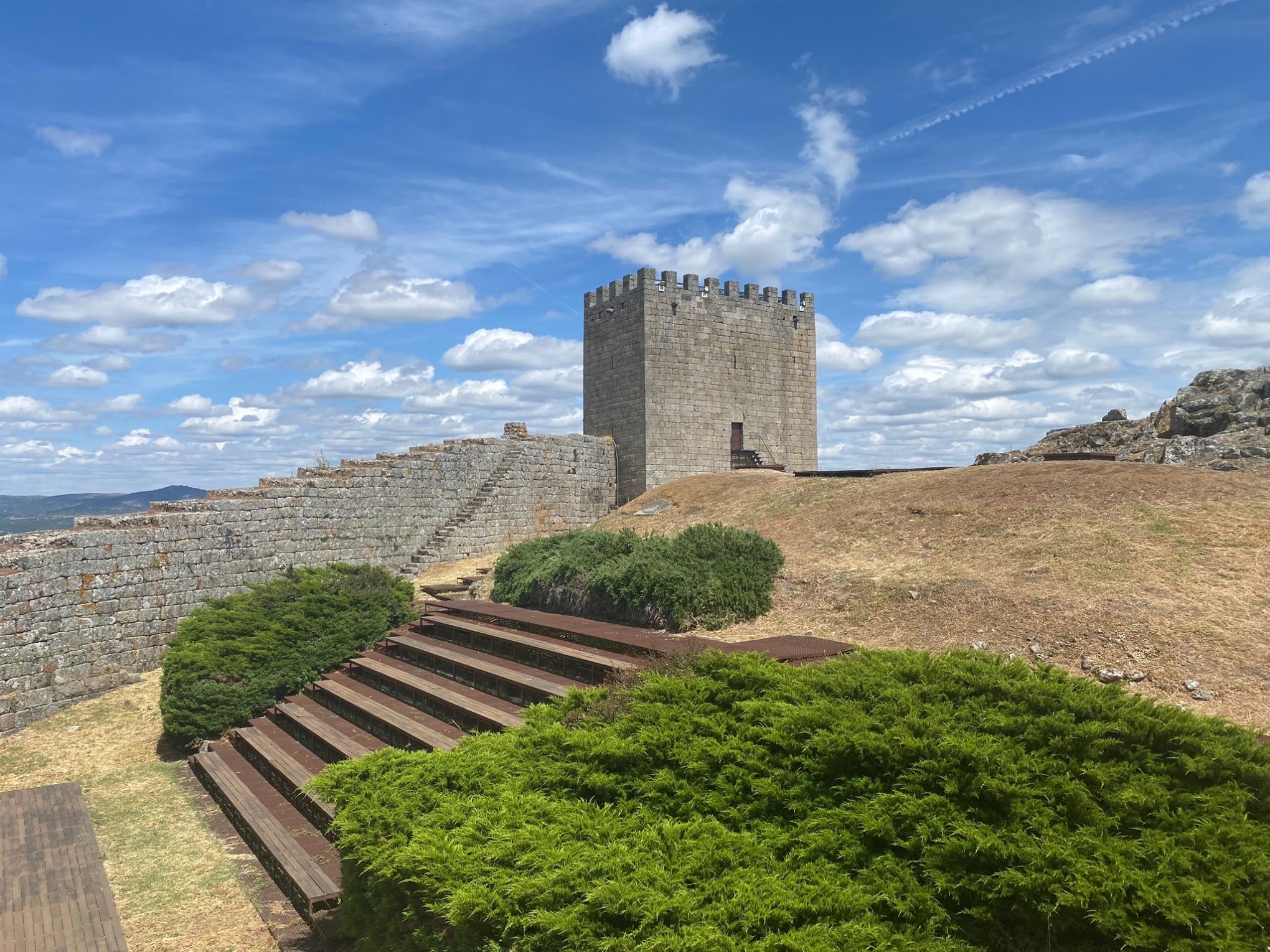 Stone tower and wall under blue sky with clouds, surrounded by grass and shrubs.