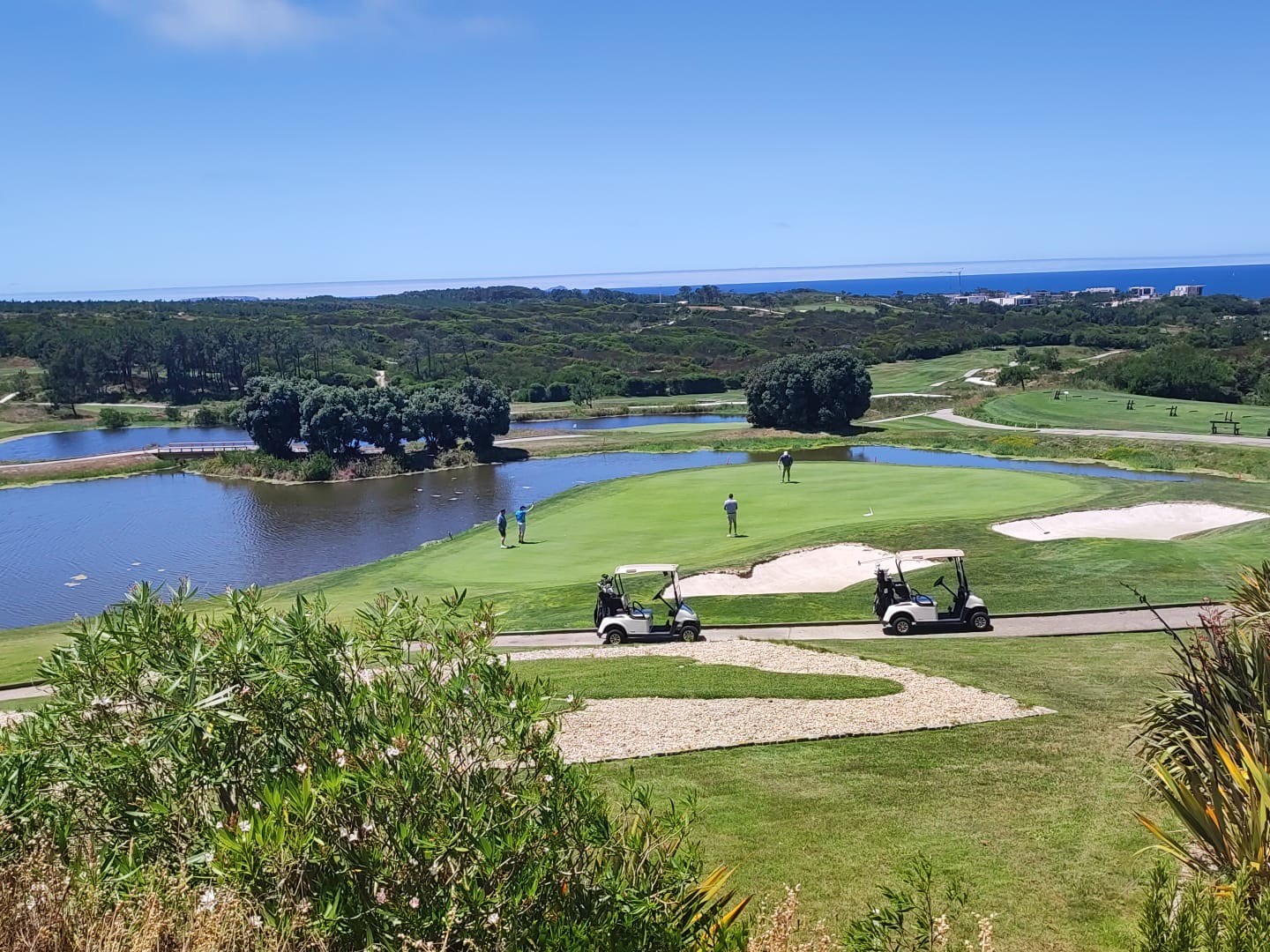 Golf course with water, golfers, and carts; ocean and blue sky in the background.