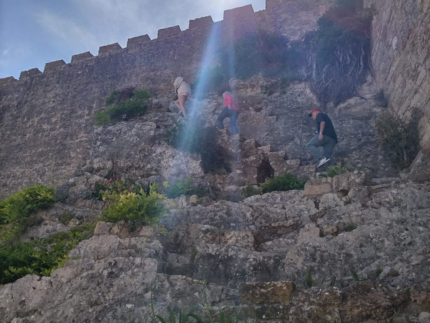 People climbing rocky steps near ancient stone wall under bright sun.