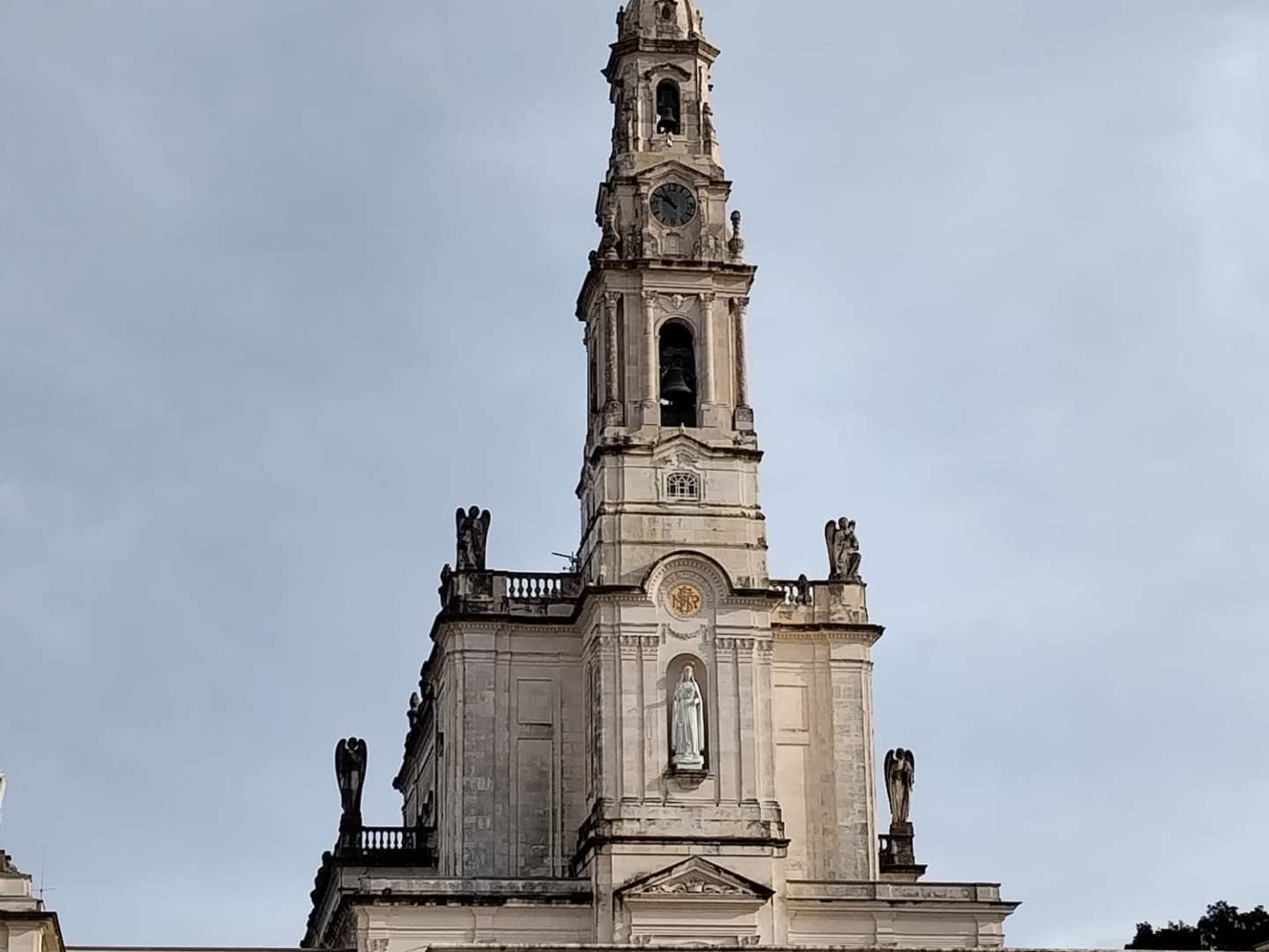 Front view of a tall church tower with a clock and cross on top, statues, and people in the foreground.